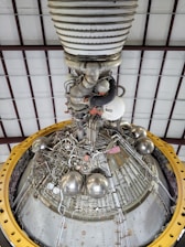 A detailed view of a complex engine or machinery component, featuring numerous metallic pipes, valves, and spherical objects. The structure is surrounded by a large yellow circular frame and is positioned against a ceiling with a grid-like pattern of beams.