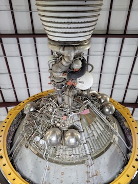 A detailed view of a complex engine or machinery component, featuring numerous metallic pipes, valves, and spherical objects. The structure is surrounded by a large yellow circular frame and is positioned against a ceiling with a grid-like pattern of beams.