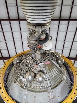 A detailed view of a complex engine or machinery component, featuring numerous metallic pipes, valves, and spherical objects. The structure is surrounded by a large yellow circular frame and is positioned against a ceiling with a grid-like pattern of beams.