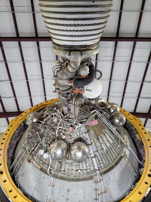 A detailed view of a complex engine or machinery component, featuring numerous metallic pipes, valves, and spherical objects. The structure is surrounded by a large yellow circular frame and is positioned against a ceiling with a grid-like pattern of beams.