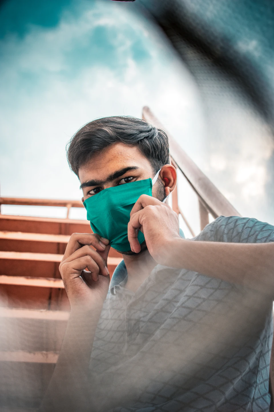 The image shows a young man wearing a green face mask, possibly for protection or to follow health guidelines during a pandemic. The man is seated on a wooden bench with his hands resting on his knees, and he appears to be looking directly at the camera.