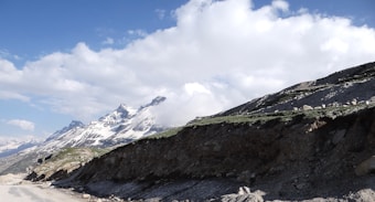 A dramatic landscape featuring snow-capped mountains under an expansive blue sky filled with fluffy clouds. The rocky terrain and a dirt road lead towards the peaks, while patches of green grass are visible on the hillside.