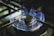 A lit gas stove burner with blue flames emanating in a symmetrical pattern around the circular metal element. The metal supports of the stovetop are visible, framing the burner, and the surrounding area shows small details typical of a kitchen stove.