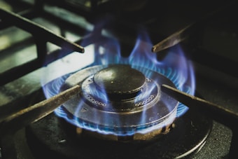 A lit gas stove burner with blue flames emanating in a symmetrical pattern around the circular metal element. The metal supports of the stovetop are visible, framing the burner, and the surrounding area shows small details typical of a kitchen stove.