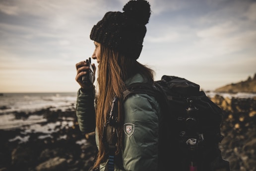 A person wearing a dark hat and a green jacket appears to be speaking into a handheld radio. They are standing near a rocky shoreline with the ocean in the background. The person has a large backpack, suggesting a hiking or outdoor adventure scene.