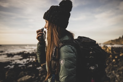 A person wearing a dark hat and a green jacket appears to be speaking into a handheld radio. They are standing near a rocky shoreline with the ocean in the background. The person has a large backpack, suggesting a hiking or outdoor adventure scene.