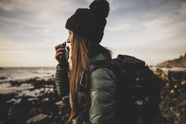 A person wearing a dark hat and a green jacket appears to be speaking into a handheld radio. They are standing near a rocky shoreline with the ocean in the background. The person has a large backpack, suggesting a hiking or outdoor adventure scene.