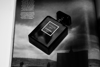 A black perfume bottle labeled 'Coco Noir Chanel Paris' is placed on an open magazine page. The page features a grayscale image of a landscape with silhouetted figures and text written in light color.