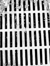A metal grate with parallel bars covers a drain or opening. The surface appears weathered with debris and leaves scattered on top, suggesting an outdoor setting.