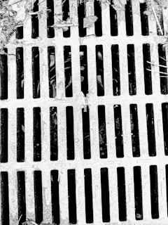 A metal grate with parallel bars covers a drain or opening. The surface appears weathered with debris and leaves scattered on top, suggesting an outdoor setting.