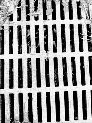 A metal grate with parallel bars covers a drain or opening. The surface appears weathered with debris and leaves scattered on top, suggesting an outdoor setting.