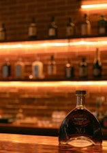A bottle of Martell XO sits prominently on a polished wooden bar counter. In the background, a brick wall with various bottles is softly illuminated by warm, ambient lighting.