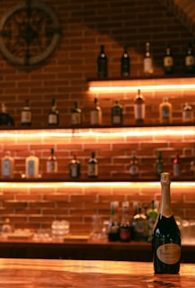 A dimly lit bar area with a brick wall background, featuring shelves filled with various bottles. A champagne bottle prominently sits on a polished wooden bar counter. Soft, warm lighting enhances the cozy and intimate atmosphere.