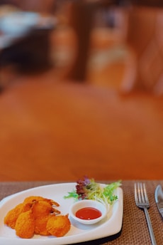 A plated dish featuring golden-brown fried shrimp alongside a small serving of colorful salad. A small white ramekin contains red dipping sauce. The plate is set on a brown woven placemat with a fork and knife visible on the side.