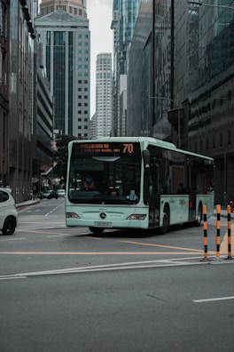 A green bus is traveling through an urban cityscape lined with tall skyscrapers and reflective glass buildings. The street is wide, and a few vehicles are visible in the background. The bus display shows route number 70.