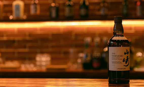 A bottle of Yamazaki single malt Japanese whisky is prominently positioned on a wooden bar counter, with an illuminated, blurred background of brick wall and bottles creating a warm and inviting atmosphere.