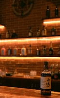 A bottle of whiskey is prominently displayed on a wooden counter in a dimly lit bar with soft amber lighting. The background shows shelves lined with various liquor bottles against a brick wall, creating a warm and inviting atmosphere.