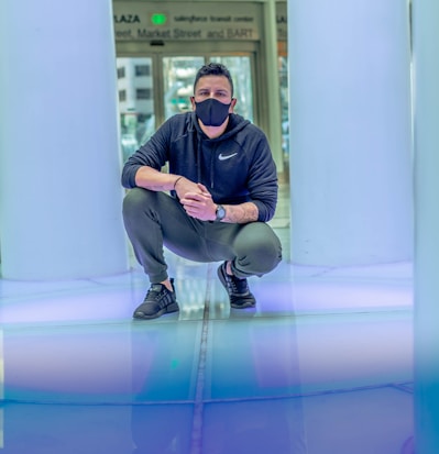 A person wearing a black mask and dark athletic clothing is crouched down in a brightly lit environment. The setting features smooth, reflective surfaces with cool tones, and a backdrop that includes signage. The overall atmosphere is modern and stylized.