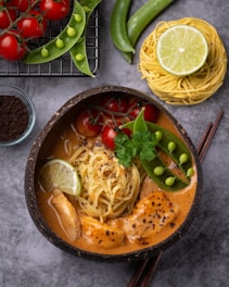 A bowl of creamy noodle soup features fresh ingredients like sliced lime, cherry tomatoes on the vine, sugar snap peas, and pieces of seasoned chicken. The soup is garnished with green cilantro leaves and black pepper. Nearby, uncooked noodles are neatly arranged with a lime half on top. Whole sugar snap peas and a wire rack holding cherry tomatoes complete the composition, placed on a gray surface.