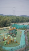 A vibrant outdoor playground with various equipment, including slides, climbing structures, and small houses. Several children and adults are enjoying the space, surrounded by lush green trees and hills in the background. An electricity pylon and power lines are visible above the trees, with a clear sky.