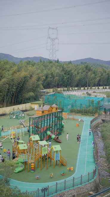 A vibrant outdoor playground with various equipment, including slides, climbing structures, and small houses. Several children and adults are enjoying the space, surrounded by lush green trees and hills in the background. An electricity pylon and power lines are visible above the trees, with a clear sky.