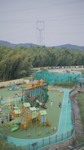 A vibrant outdoor playground with various equipment, including slides, climbing structures, and small houses. Several children and adults are enjoying the space, surrounded by lush green trees and hills in the background. An electricity pylon and power lines are visible above the trees, with a clear sky.