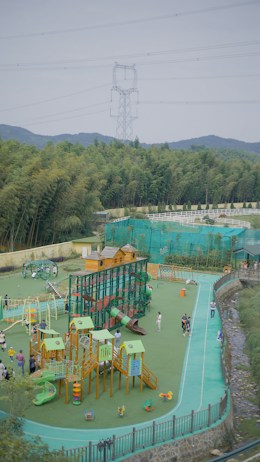 A vibrant outdoor playground with various equipment, including slides, climbing structures, and small houses. Several children and adults are enjoying the space, surrounded by lush green trees and hills in the background. An electricity pylon and power lines are visible above the trees, with a clear sky.
