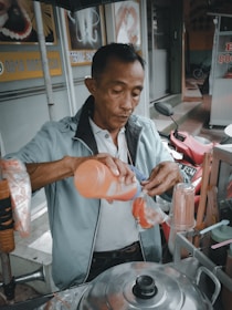 A man is preparing a drink at a street food stall. He is holding an orange bottle, pouring the contents into a plastic bag filled with ice. There are snacks and utensils around him as he works at his stall, with a scooter in the background.