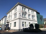 A multi-story white building with classic architectural details such as decorative window frames and a corner design. It appears to be a bank, as indicated by a sign on the top floor. In front of the building, there's a streetlight and a street sign, along with some shrubs and a tree. The sky is clear, suggesting good weather.