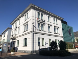 A multi-story white building with classic architectural details such as decorative window frames and a corner design. It appears to be a bank, as indicated by a sign on the top floor. In front of the building, there's a streetlight and a street sign, along with some shrubs and a tree. The sky is clear, suggesting good weather.