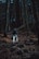 A person wearing a backpack and outdoor gear is walking through a dense forest filled with tall, bare trees and scattered leaves on the ground. The scene suggests a late autumn or early winter setting with low light filtering through the tree canopy.