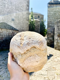 A hand holds a large, round loaf of rustic bread against a backdrop of old stone buildings. The texture of the bread is rough with a light dusting of flour, and it appears freshly baked. The sun casts bright, natural light on the scene, creating a warm and inviting atmosphere.