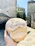 A hand holds a large, round loaf of rustic bread against a backdrop of old stone buildings. The texture of the bread is rough with a light dusting of flour, and it appears freshly baked. The sun casts bright, natural light on the scene, creating a warm and inviting atmosphere.