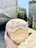 A hand holds a large, round loaf of rustic bread against a backdrop of old stone buildings. The texture of the bread is rough with a light dusting of flour, and it appears freshly baked. The sun casts bright, natural light on the scene, creating a warm and inviting atmosphere.
