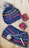 A knitted hat with a pom-pom and a matching cowl are displayed on a light wooden surface. Both pieces feature colorful stripes including blue, pink, yellow, and green. Alongside them are two crocheted squares and a ball of yarn with a crochet hook.