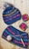 A knitted hat with a pom-pom and a matching cowl are displayed on a light wooden surface. Both pieces feature colorful stripes including blue, pink, yellow, and green. Alongside them are two crocheted squares and a ball of yarn with a crochet hook.