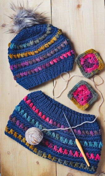 A knitted hat with a pom-pom and a matching cowl are displayed on a light wooden surface. Both pieces feature colorful stripes including blue, pink, yellow, and green. Alongside them are two crocheted squares and a ball of yarn with a crochet hook.
