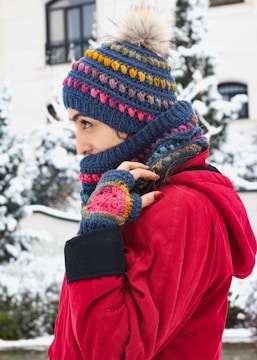 A person is standing outdoors wearing winter clothing, including a red coat and a crocheted hat with a pom-pom, scarf, and fingerless gloves, all featuring colorful patterns. The background shows a snowy landscape with trees and a building.