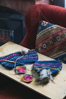 A wooden table displays several colorful knitted items along with crochet squares and a ball of yarn. A crochet hook is placed among the items. The background features a textured, multi-colored cushion and a red upholstered chair.