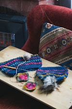 A wooden table displays several colorful knitted items along with crochet squares and a ball of yarn. A crochet hook is placed among the items. The background features a textured, multi-colored cushion and a red upholstered chair.
