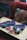 A wooden table displays several colorful knitted items along with crochet squares and a ball of yarn. A crochet hook is placed among the items. The background features a textured, multi-colored cushion and a red upholstered chair.