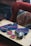 A wooden table displays several colorful knitted items along with crochet squares and a ball of yarn. A crochet hook is placed among the items. The background features a textured, multi-colored cushion and a red upholstered chair.