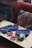 A wooden table displays several colorful knitted items along with crochet squares and a ball of yarn. A crochet hook is placed among the items. The background features a textured, multi-colored cushion and a red upholstered chair.