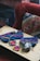 A wooden table displays several colorful knitted items along with crochet squares and a ball of yarn. A crochet hook is placed among the items. The background features a textured, multi-colored cushion and a red upholstered chair.