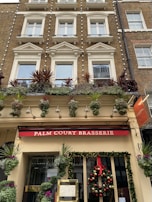 A multi-story building facade with classic architecture features, including large windows with decorative moldings. The first level displays a restaurant sign reading 'Palm Court Brasserie' in white letters on a red background. It is adorned with greenery and festive decorations, including a wreath with red ribbon and ornaments. Potted plants with colorful foliage are arranged on the ground floor and along the second floor balcony.