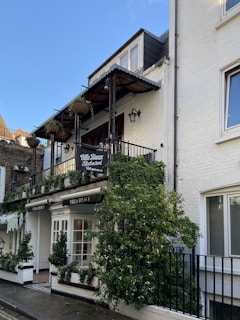 A two-story building with a white exterior and a black metal balcony. The facade is adorned with lush green plants, and the entrance features glass doors with an awning above. Hanging planters and wall-mounted lamps add to the rustic charm. A sign reads 'Villa Bianca Restaurant'.