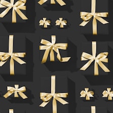 A pattern of variously sized black gift boxes adorned with shiny golden ribbons, arranged neatly against a dark background. The lighting casts subtle shadows, highlighting the gleaming texture of the ribbons.
