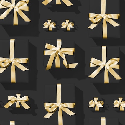 A pattern of variously sized black gift boxes adorned with shiny golden ribbons, arranged neatly against a dark background. The lighting casts subtle shadows, highlighting the gleaming texture of the ribbons.
