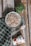A bowl of yogurt granola with nuts and dried fruits sits on a cooling rack, accompanied by a spoon with some yogurt on it. Next to the setup, there is a can of Alpro strawberry yogurt. A checkered cloth and a small green plant add to the rustic presentation, all placed on a wooden surface.