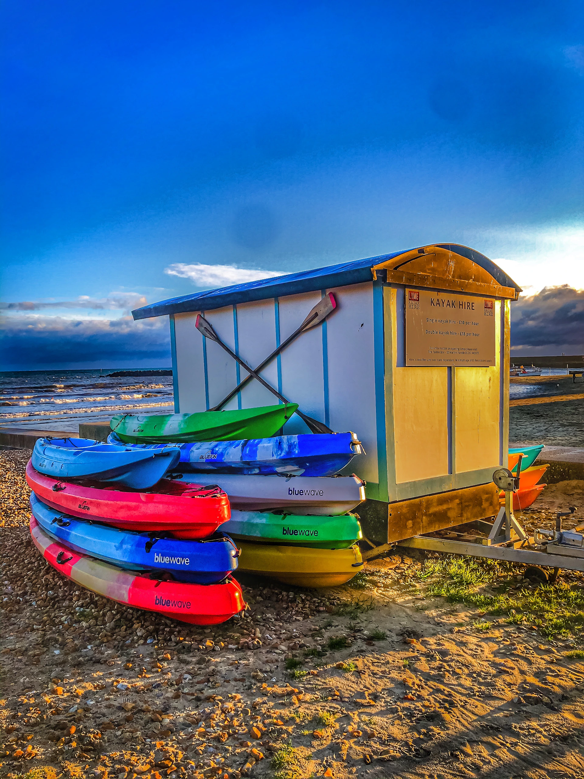 Several colorful kayaks are stacked next to a small wooden hut on a pebbled beach, with the sea and a dramatic blue sky with some clouds in the background. The hut has a sign indicating kayak rentals.